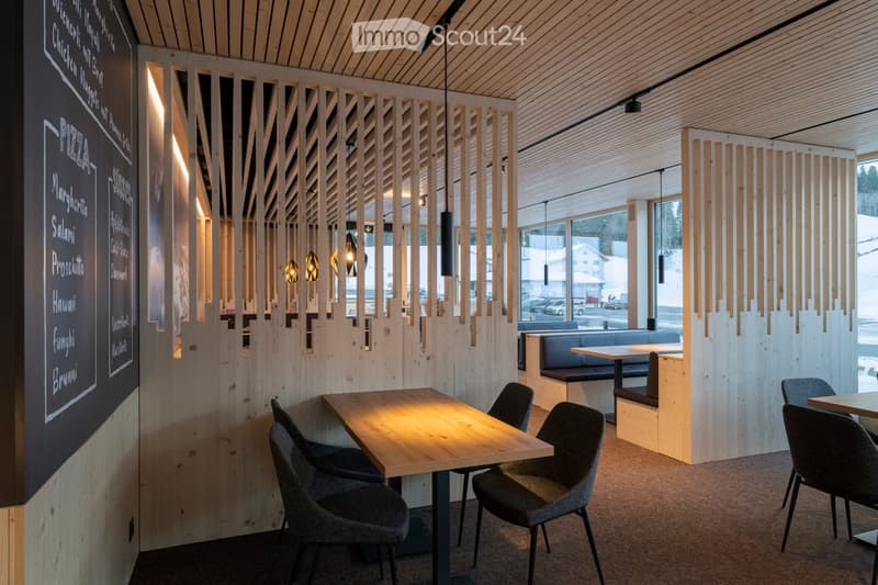 Modern restaurant interior with wooden tables, grey chairs, and a bench seating area. A menu board is visible on the left side.
