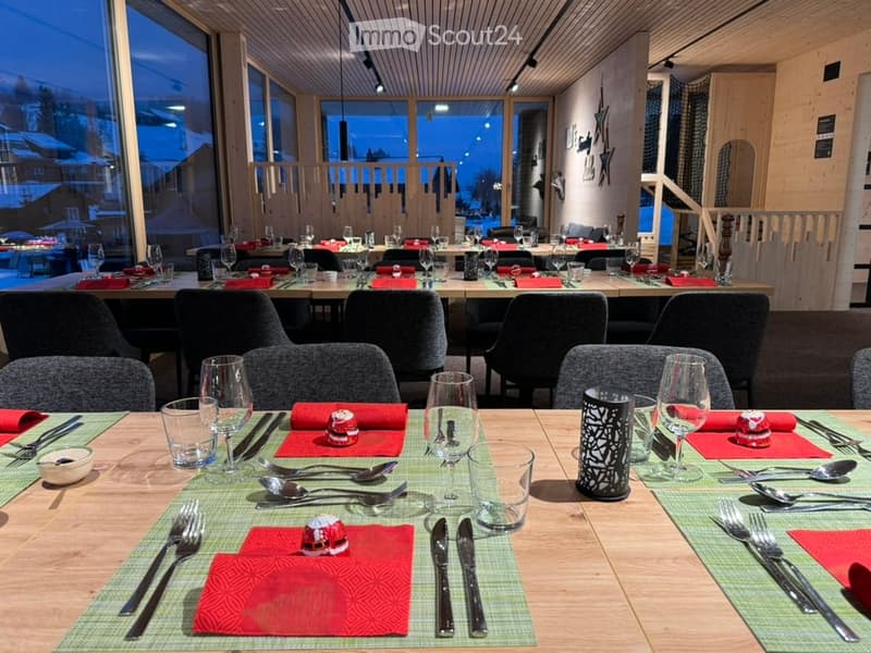 A restaurant or dining area with long wooden tables, chairs, and place settings including wine glasses, cutlery, and napkins. Large windows offer a view of the snowy outdoor scene.