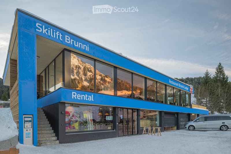 Modern ski rental building with large blue signage reading "Skilift Brunnli" and "Rental". Features large windows, a staircase, and a parked van in front.