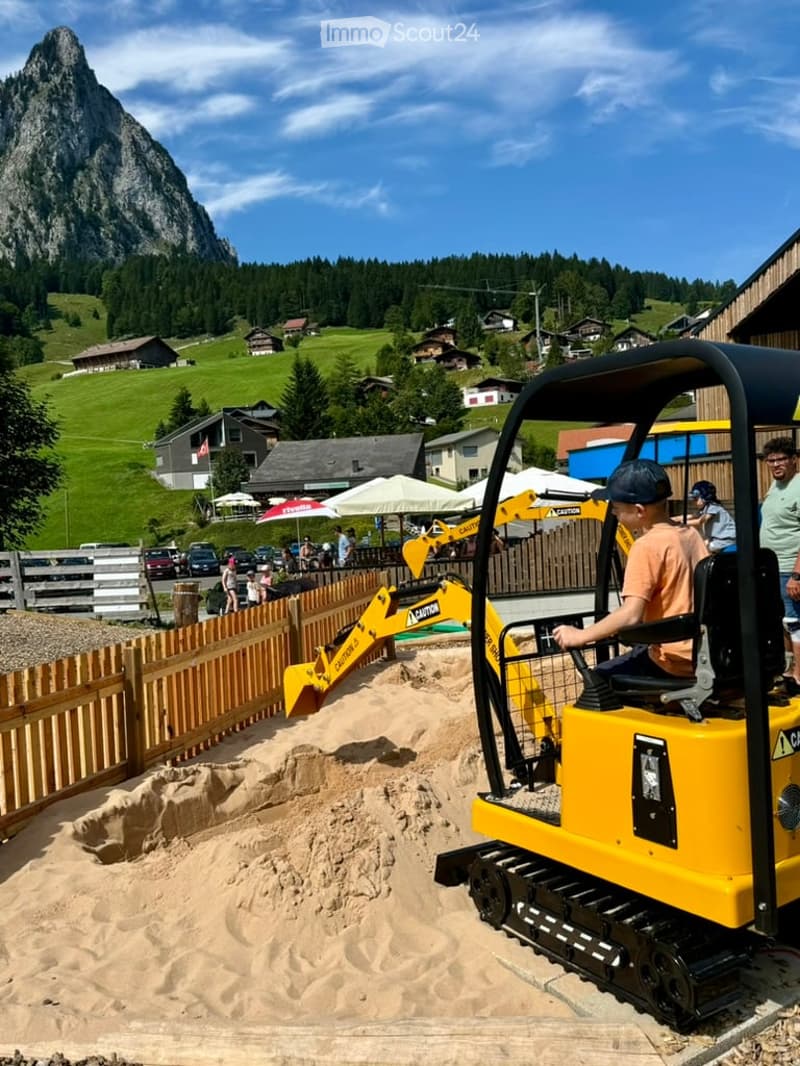 Outdoor area with a young boy operating a small yellow excavator in a sandy pit, surrounded by wooden fences, other children, and a scenic mountain backdrop with houses and green fields.