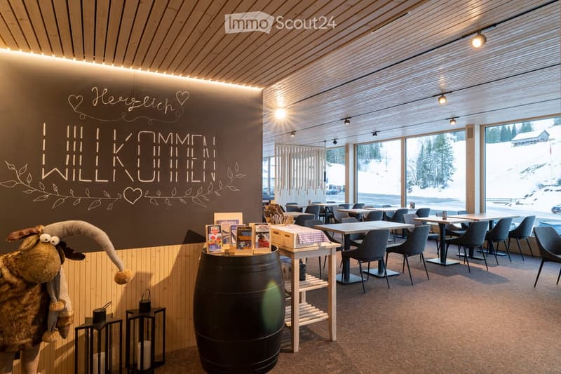 Modern restaurant interior with wooden ceiling, large windows showing snowy landscape, tables and chairs, barrel centerpiece, decorative shelf, and plush animal figure.