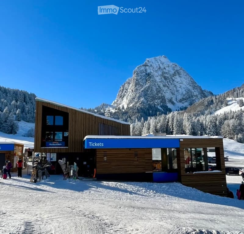 Modern wooden building with a ticket booth and ski equipment storage, situated in a snowy mountain landscape with a prominent peak in the background.