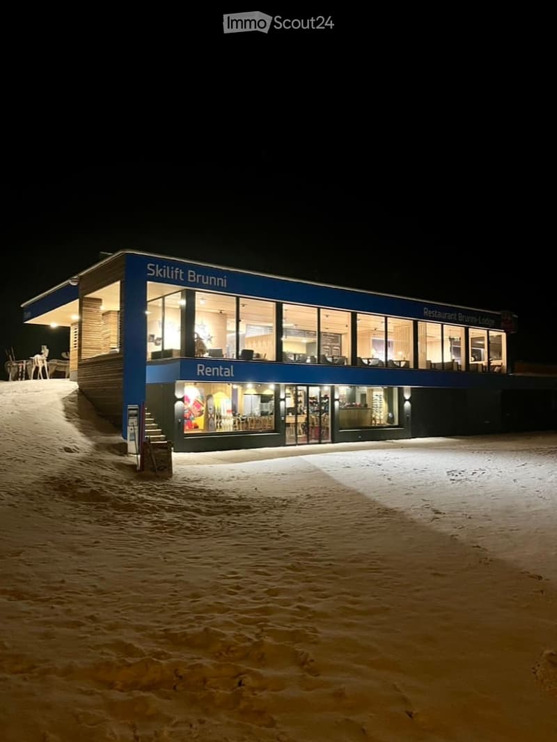 Nighttime view of a modern building labeled "Skilift Bruni" with a restaurant and rental area, illuminated by exterior lighting, surrounded by snow.