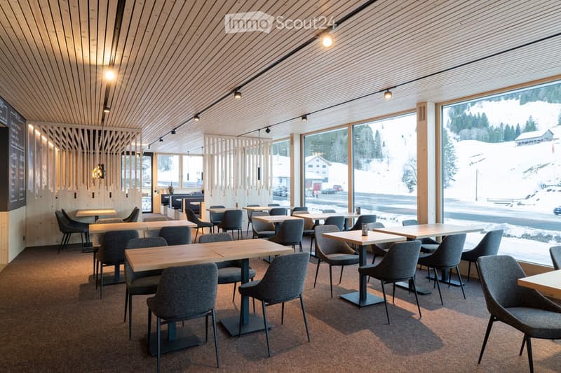 Modern restaurant interior with wooden tables, grey chairs, large windows overlooking snowy landscape, warm lighting.