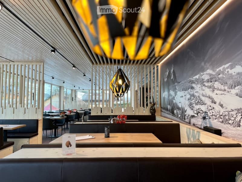 Modern restaurant interior with wooden tables, blue upholstered benches, geometric pendant lights, and a large mural of a snowy mountain landscape.