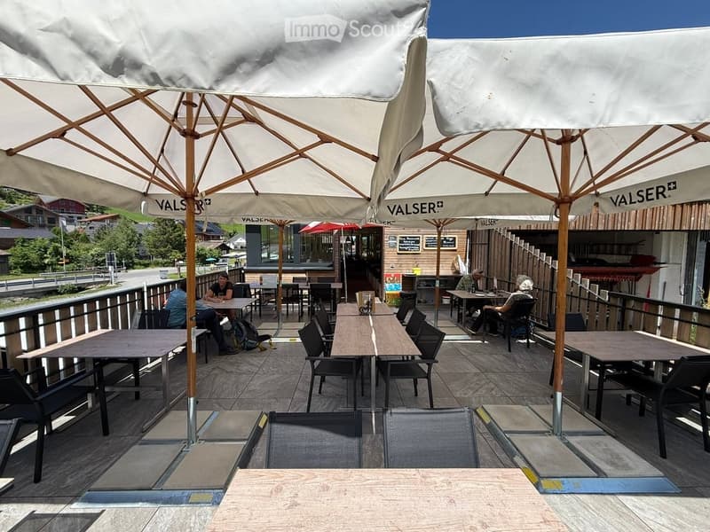 Outdoor seating area with tables, chairs, and large umbrellas, surrounded by a wooden deck and a scenic mountain backdrop.