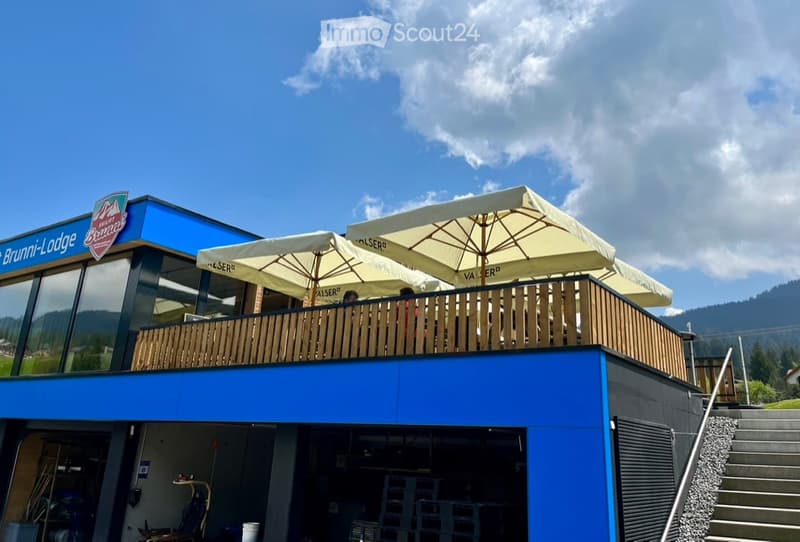 Modern building with a large balcony area featuring umbrellas and seating, surrounded by a scenic mountain landscape.