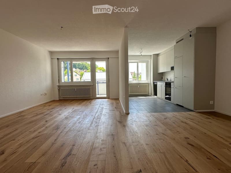 Empty room with wooden flooring, large windows, and a view of the outside. The adjacent kitchen area has white cabinets and a tiled floor.