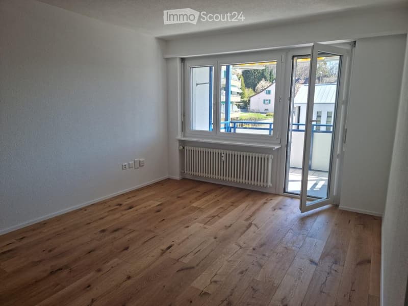 Empty room with wooden flooring, white walls, a radiator, and a large window with a sliding glass door leading to a balcony.
