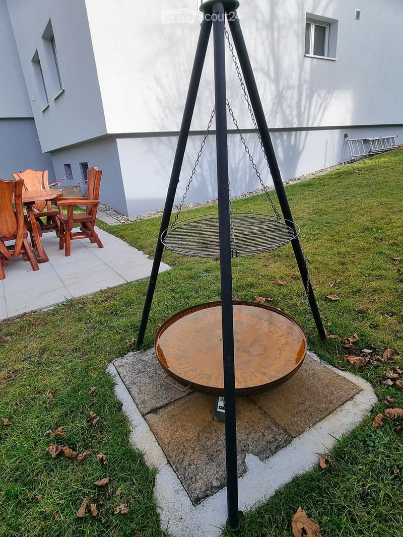 Garden area with a metal fire pit, wooden table, and chairs, surrounded by grass and a paved pathway.