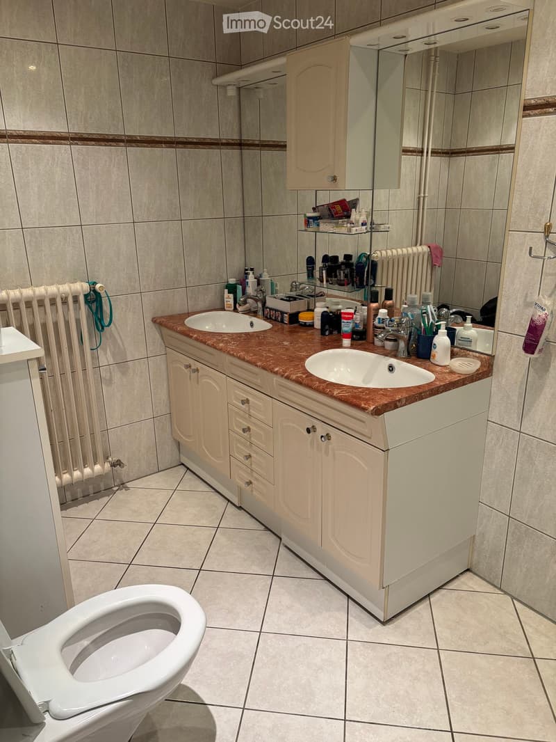 Bathroom with a double sink vanity, toilet, tiled walls, and various toiletries on the counter.