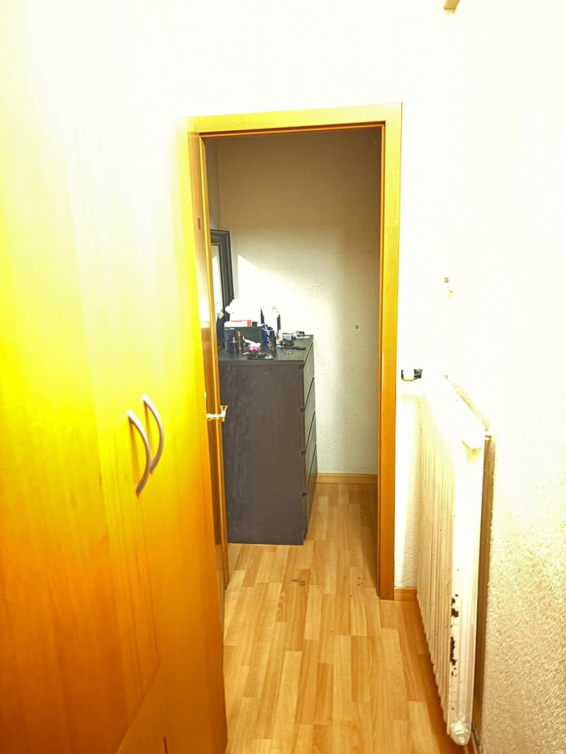 A narrow hallway with wooden flooring and white walls, leading to a small bathroom with a dark vanity and various toiletries.