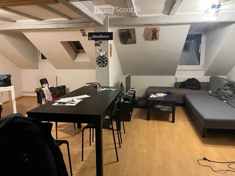 A multi-functional room with a sofa, coffee table, desk, and laptop, featuring wooden flooring and slanted ceilings.