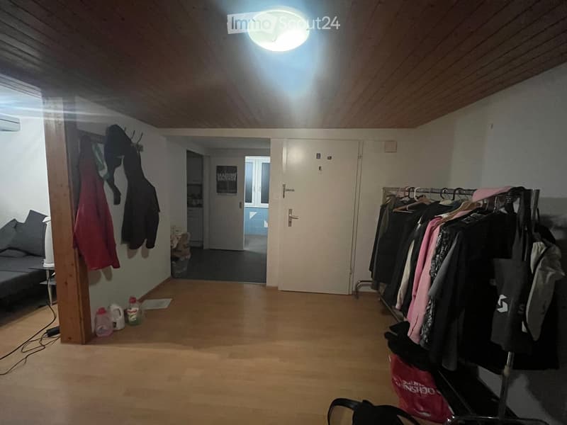 The image shows a furnished room with a wooden floor, a sofa on the left side, a clothing rack on the right side, and a doorway leading to another room. There are several bottles and a handbag visible on the floor.