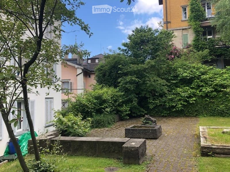 A small, enclosed garden area with gravel pathways, stone benches, and lush greenery, surrounded by residential buildings.