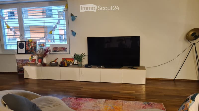 Modern living room with large windows, white cabinetry, a flat-screen TV, a floor lamp, and decorative items on the cabinet.