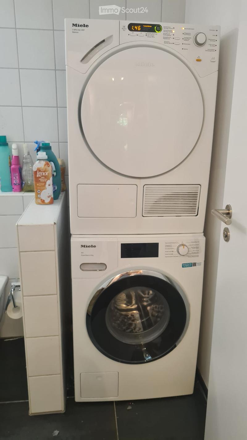 A bathroom with a stacked Miele washing machine and dryer unit, surrounded by cleaning supplies on a shelf.