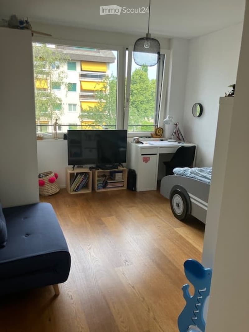 A multi-functional room with a sofa, TV, desk, chair, bookshelf, and a view of a building outside the window.