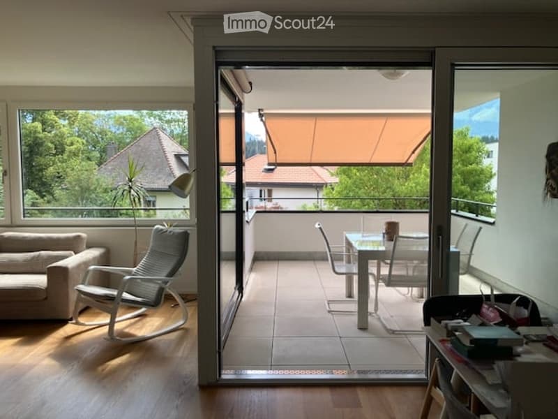 Bright living room with large windows and sliding glass doors leading to a balcony. Features a sofa, armchair, dining table with chairs, and a small shelf with books.