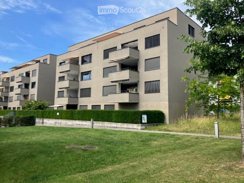 Modern multi-story residential building with balconies, surrounded by a well-maintained lawn and trees.