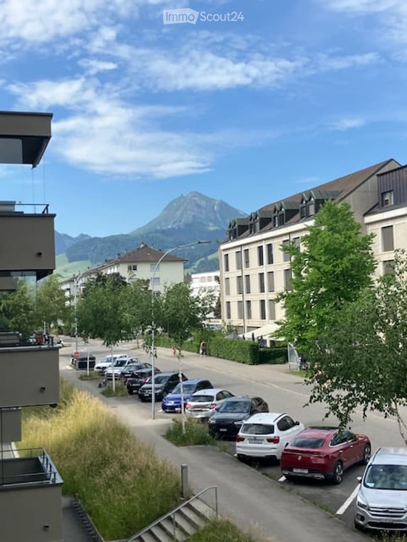 View from a balcony showing a street lined with parked cars, buildings, and a scenic mountain backdrop.