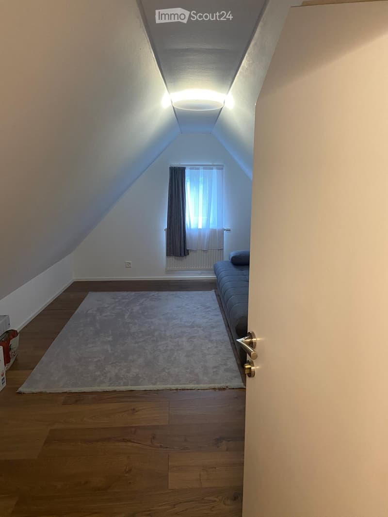 Attic room with wooden flooring, slanted ceilings, and a small window with curtains. Features a sofa and a rug.