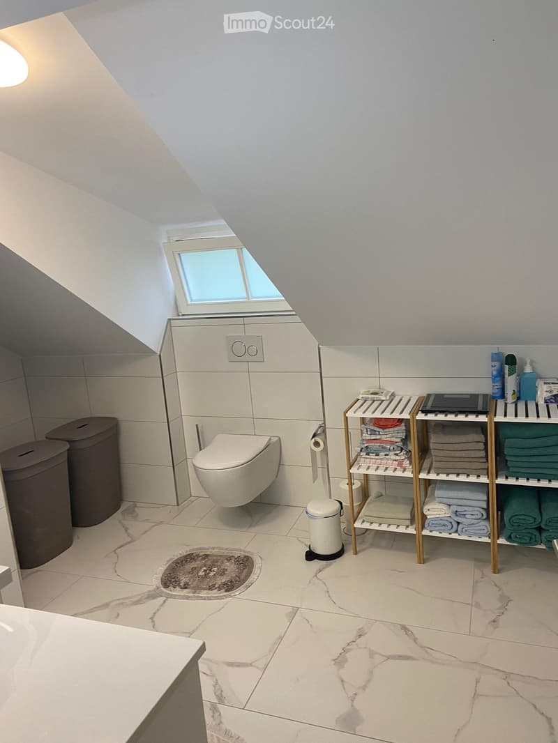 Bathroom with white tiles, white toilet, white sink, wooden shelves with towels, trash bins, and a window