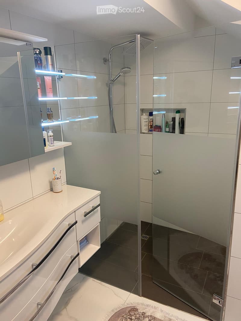 modern bathroom with white tiles, glass shower enclosure, white vanity with drawers, shelves with toiletries, and a rug