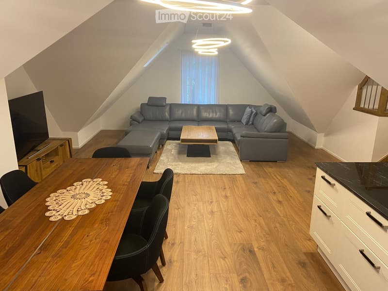 A modern living room with a gray leather sectional sofa, a wooden coffee table, a wooden dining table, black chairs, a flat-screen TV, a wooden entertainment center, a beige rug, and a wooden staircase.