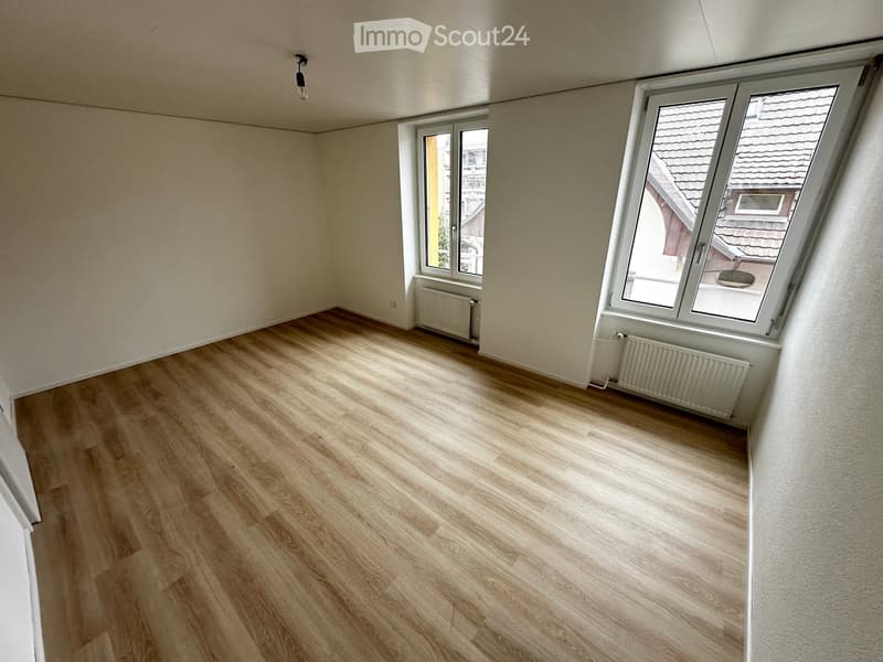 Empty room with wooden flooring, white walls, and large windows offering a view of the roof and surrounding buildings.