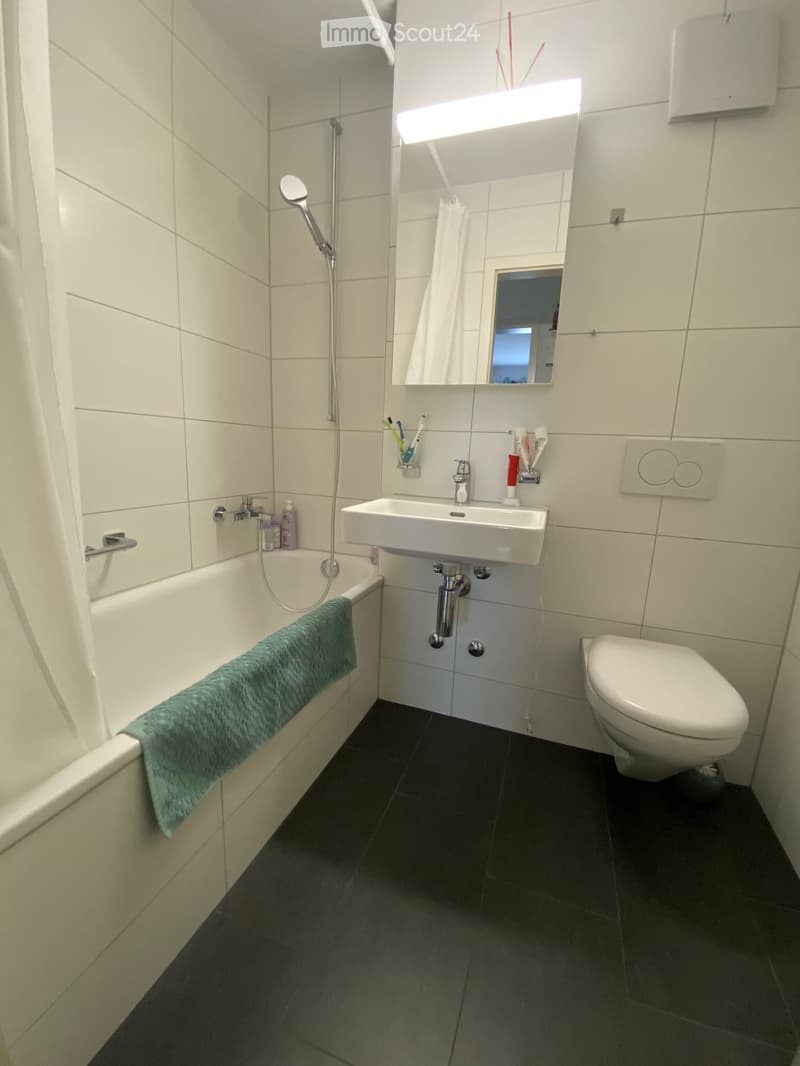 Modern bathroom with white tiles, bathtub, toilet, sink, mirror, and a green towel on the edge of the bathtub.
