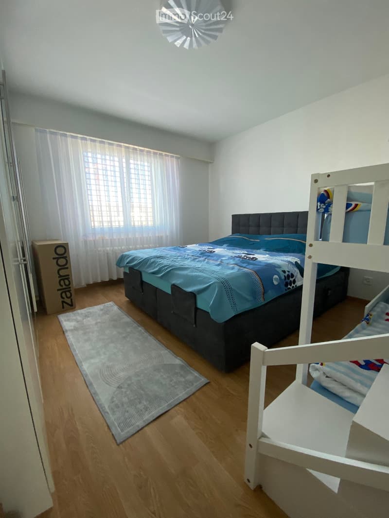 Bright bedroom with wooden flooring, a double bed with blue bedding, a bunk bed, a grey rug, and white curtains on the window.