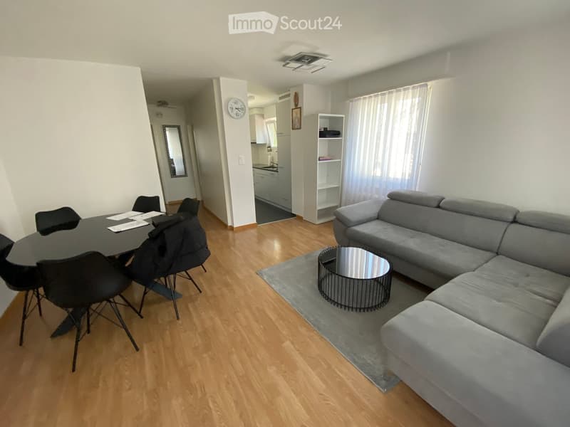 Modern living room with grey sectional sofa, round coffee table, dining table with chairs, and a small kitchen area visible in the background.