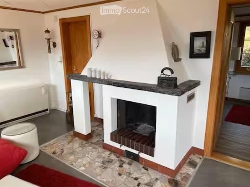 Cozy living room with a central fireplace, a white stool, a mirror on the wall, and a picture frame. The room features wooden doors and a slanted ceiling.