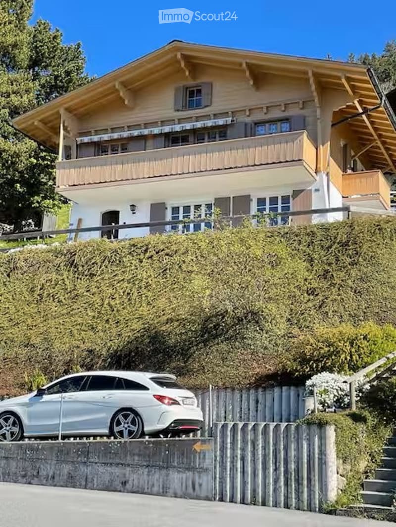 Two-story wooden house with balconies, surrounded by greenery, with a parked car in front.