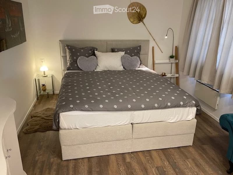 Cozy bedroom with a large bed, grey and white bedding, nightstand with lamp, and a small chair near the window.