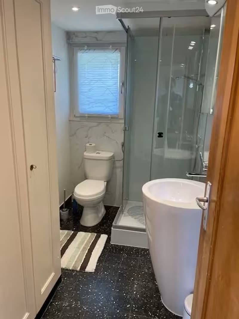 Modern bathroom with a large, freestanding sink, toilet, and a glass-enclosed shower. The walls feature marble tiles, and the floor is black with a striped bath mat.