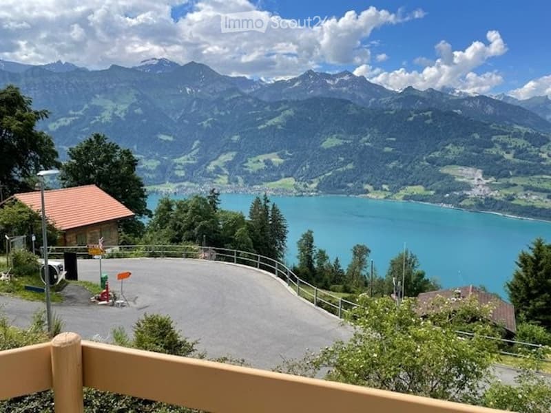 Balcony overlooking a scenic lake and mountain range, with a small structure and parking area visible below.