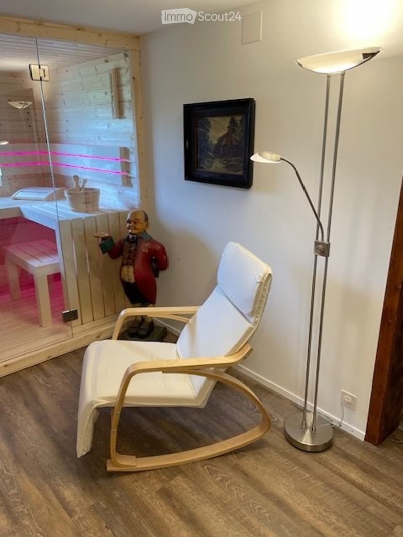 A modern, minimalist room with a white rocking chair, a tall floor lamp, a framed picture on the wall, and a small decorative statue near a glass-enclosed sauna area.