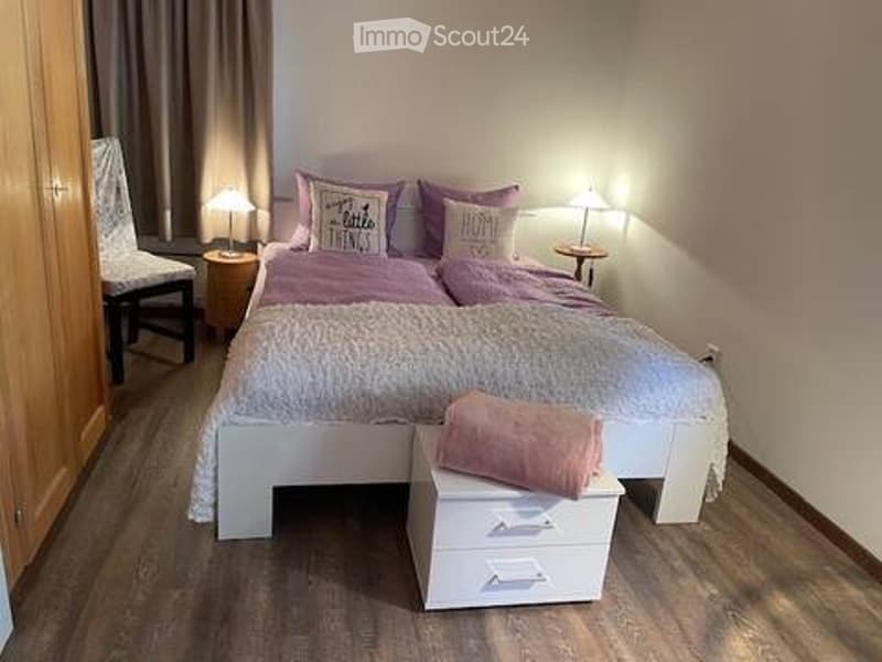 Cozy bedroom with a white bed, pink pillows and blankets, two nightstands with lamps, a wooden chair, and a small drawer with a pink towel on top.