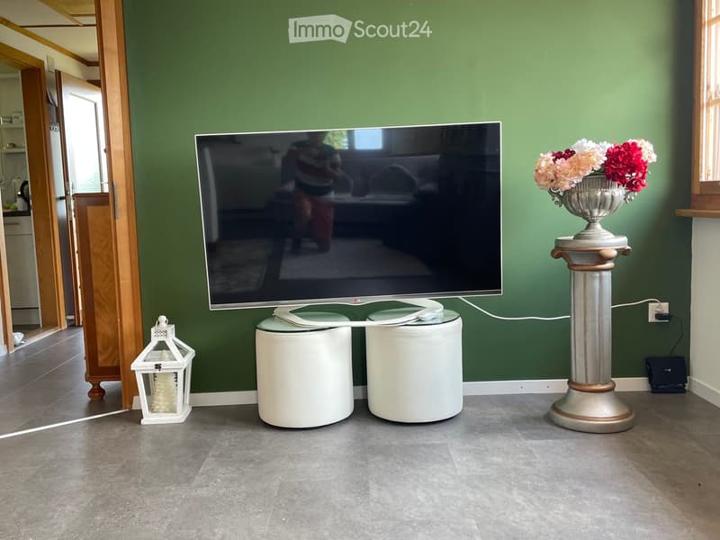 Living room with green walls, a large flat-screen TV on a white table, a decorative vase with flowers, and a lantern on the floor.