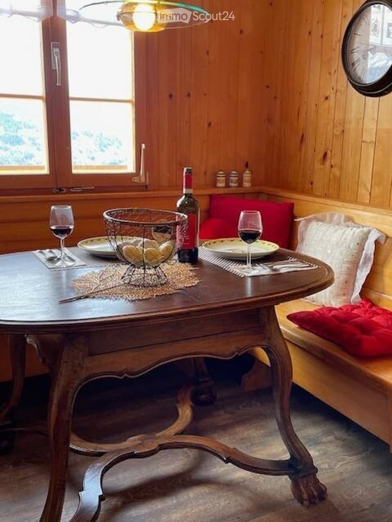 A cozy wooden-paneled dining area with a round wooden table set for two, wine glasses, a bottle of wine, and a basket of fruit. A bench with red cushions is nearby.