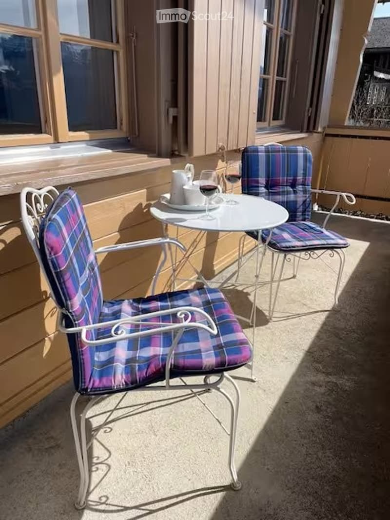 Balcony with two metal chairs and a small table, set against a yellow wall with wooden shutters and windows.
