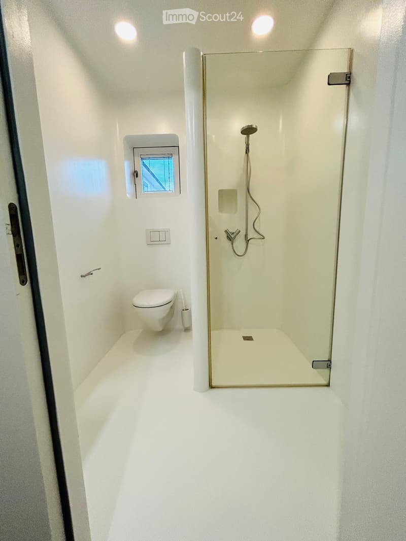 Modern, compact bathroom with white walls and floor, featuring a toilet, a glass-enclosed shower with a handheld showerhead, and a small window with blinds.