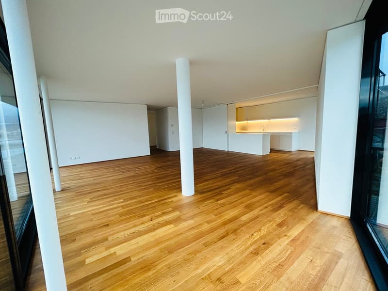 Open-plan unfurnished room with light wooden flooring, white walls, and a small kitchen area in the background.