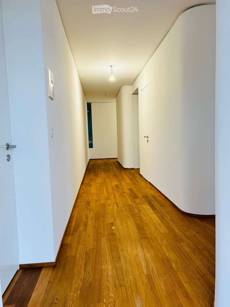 A narrow hallway with wooden flooring, white walls, and a single light fixture hanging from the ceiling. Doors are visible on both sides.