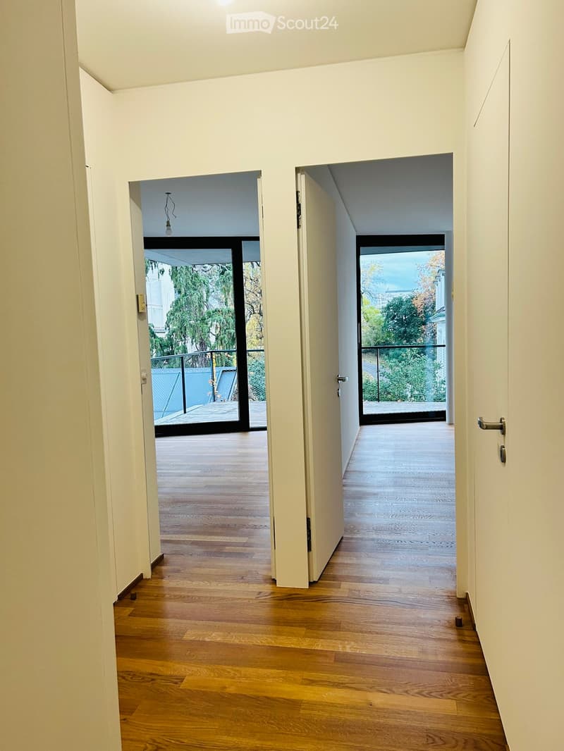 A modern hallway with light-colored wooden flooring, white walls, and two open doors leading to outdoor spaces with wooden decks and greenery visible beyond.