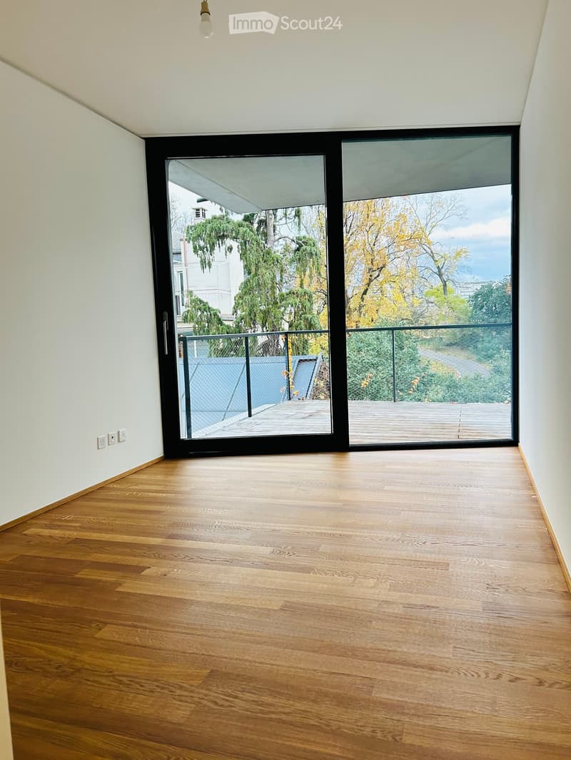 Empty room with wooden flooring, white walls, and large sliding glass doors leading to a balcony with a view of trees and a cityscape.