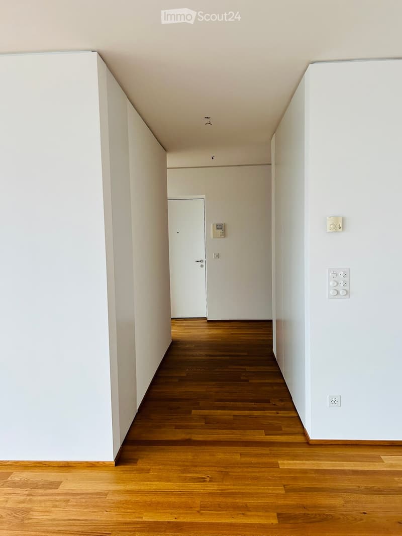 Empty hallway with wooden flooring and white walls, featuring a closed door at the end and electrical outlets on the right wall.