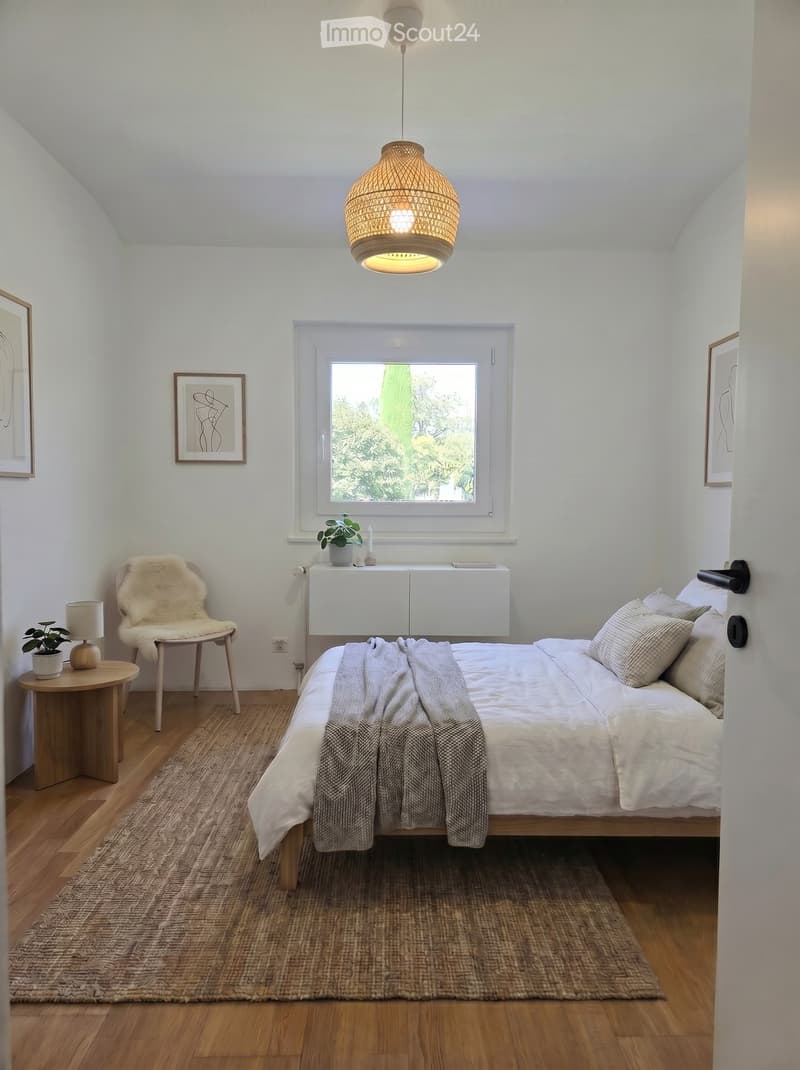 Cozy bedroom with a bed, a fur-covered chair, a small round table with a lamp, a hanging woven light fixture, and a window with a view of greenery.